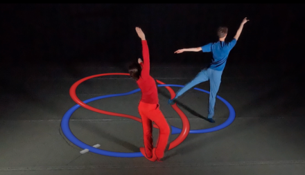 Two dancers in red and blue outfits perform with matching ribbon trails on the floor, creating interwoven loops under dramatic lighting.